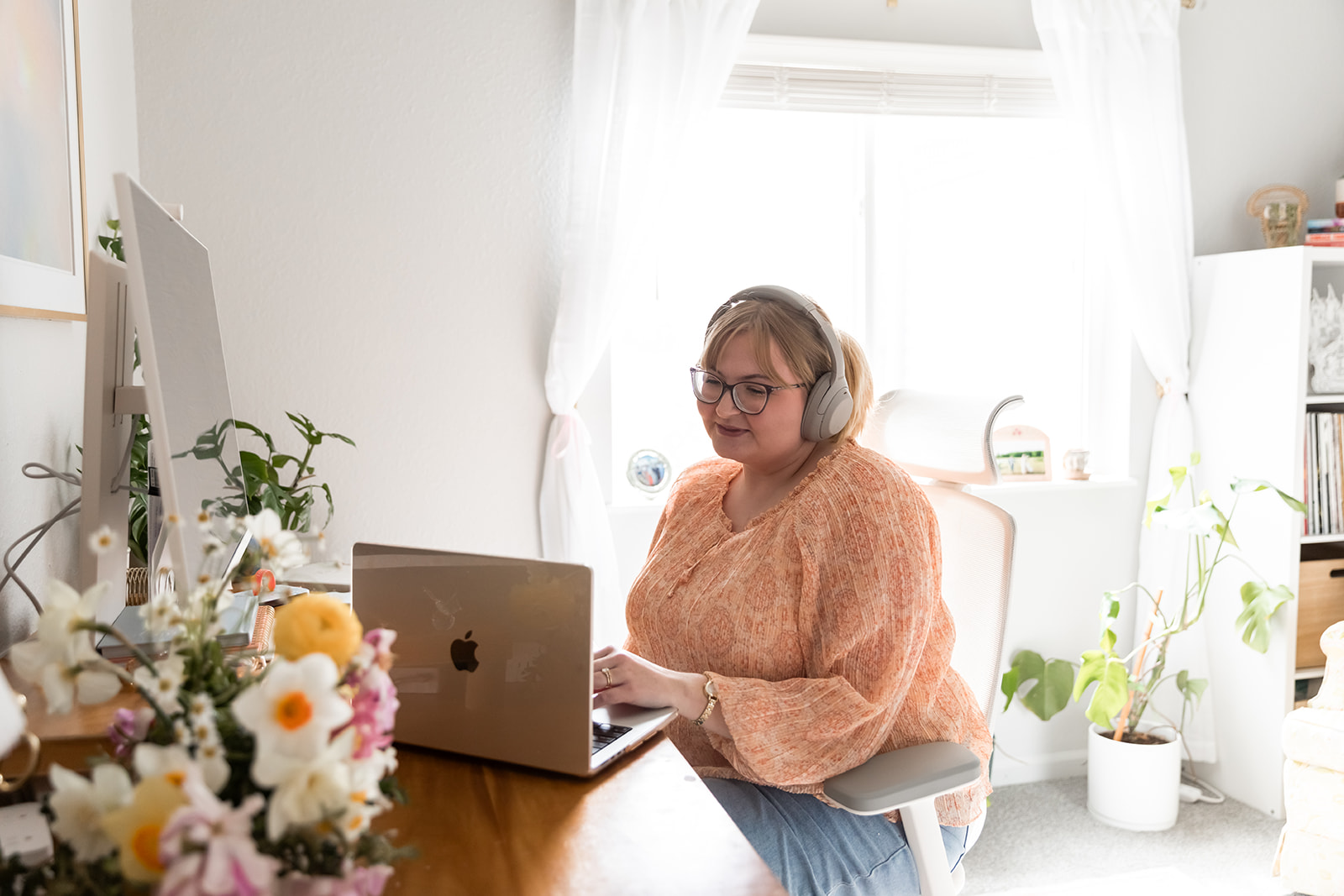 woman with blonde hair wearing pink blouse sitting at desk with computer on meeting call