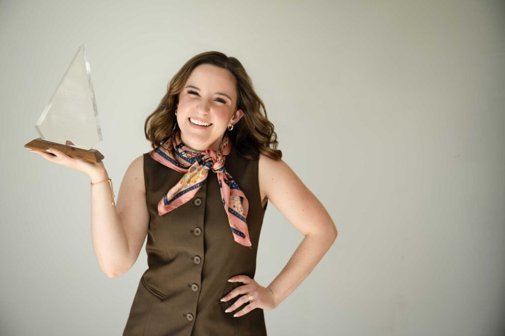 Woman with short brown hair wearing neutral green pant suit and patterned scarf holding glass award 