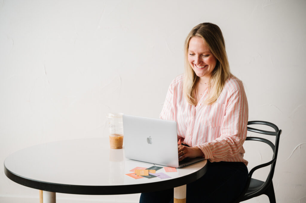 Woman with blonde hair wearing pink sweater sitting at table with silver laptop
