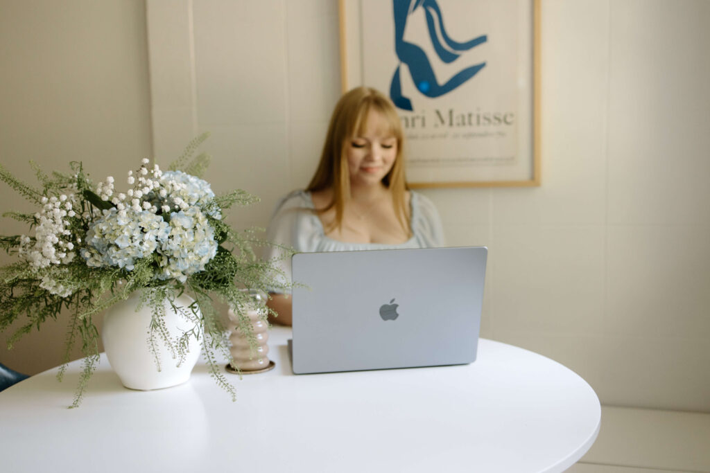 Woman with blonde hair sitting at white table with laptop typing on computer